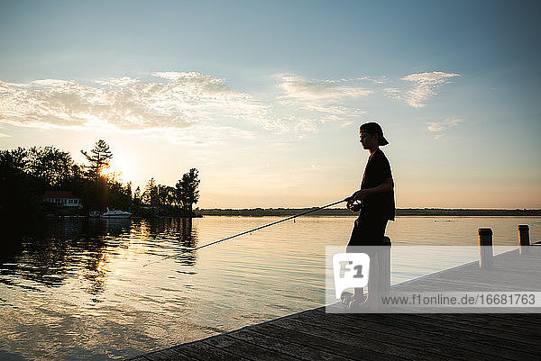 Adolescent boy fishing off dock on lake at sunset in Ontario  Canada.