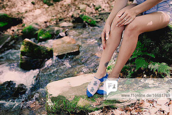 junges Hipster-Mädchen sitzt auf einem Felsen am Fluss im Wald in bedruckten hellen Socken
