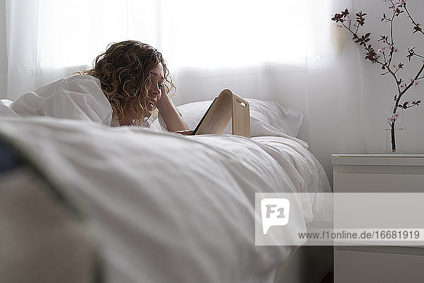 Frau mit einem Tablet-Computer auf dem Bett liegend in einem weißen Schlafzimmer.