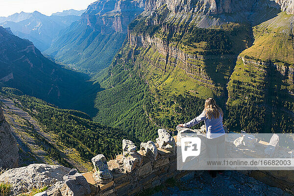 eine Frau von hinten auf einem Balkon des Nationalparks Ordesa  die von der Spitze des Berges aus das ganze Tal  die grünen Wälder  die Felsen  die Hügel und den Horizont bewundert. Horizontales Foto