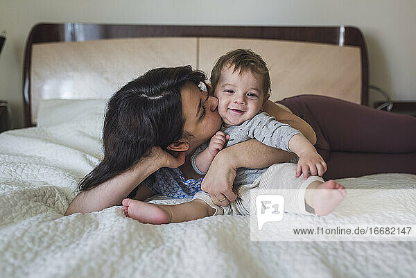 Pretty mom hugging and kissing happy year old baby on white bedspread