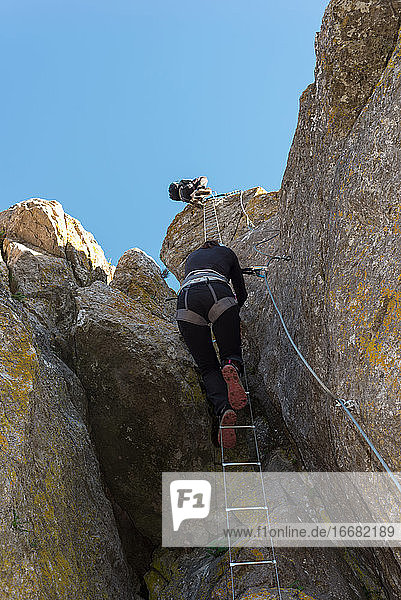 Concept: adventure. Climber woman with helmet and harness. Climbing up an iron ladder anchored to the natural wall. Via ferrata in the mountains.