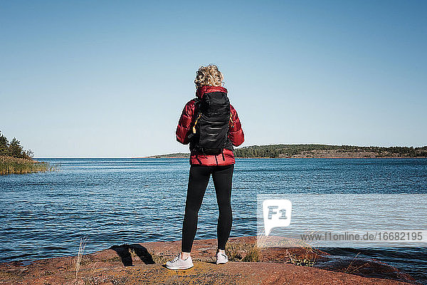Frau  die mit einem Rucksack auf einem Felsen sitzt und auf das Meer und die Inseln schaut