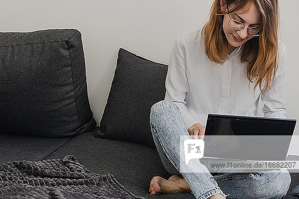 Attractive Young Woman Working from Home - Female Entrepreneur Sitting on sofa with Laptop Computer and Checking Cell Phone from Comfort of Home