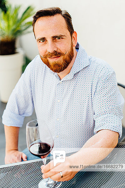 Rothaariger Mann mit Schnurrbart sitzt am Tisch mit einem Glas Rotwein