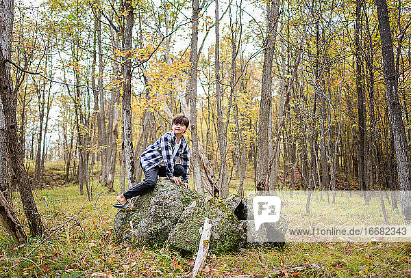 Young boy climbing on a large rock in the woods on a fall day.