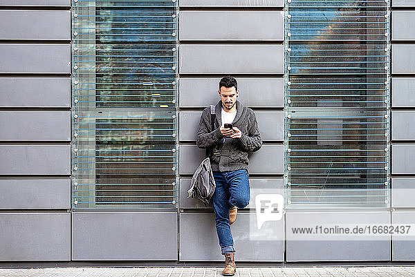 Vorderansicht eines bärtigen Mannes  der ein Telefon benutzt und sich an die Wand eines Bürogebäudes lehnt