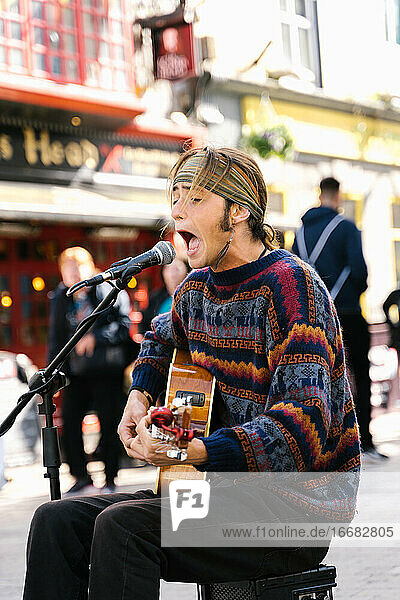 Vertical photo of a young man singing while playing guitar in the street