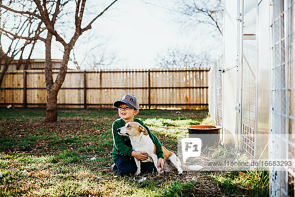 Junger Junge umarmt Corgi-Hund  während er vor einem Gewächshaus im Hinterhof sitzt
