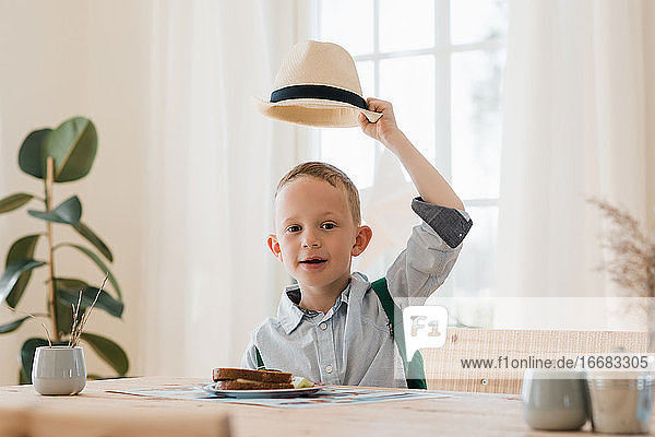Junge hält seinen Hut hoch  während er zu Hause lächelnd sein Mittagessen isst