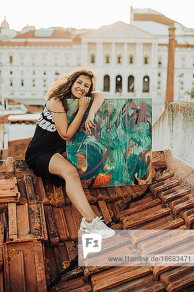 Frau umarmt ihr Kunstwerk mit glücklichem Gesicht  während sie auf einem Ziegeldach sitzt