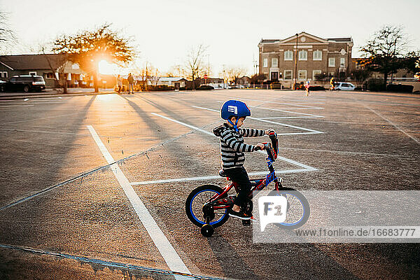 Kleiner Junge fährt Fahrrad mit Stützrädern auf einem Parkplatz bei Sonnenuntergang