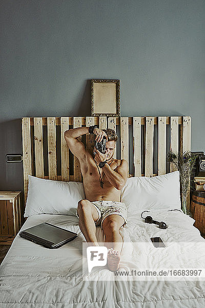 Ein Mann macht ein Foto  während er mit seinem Handy und seinem Laptop in einem Hotelbett arbeitet.
