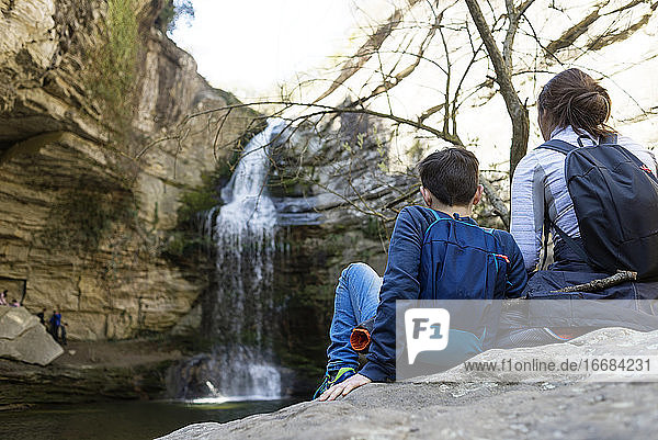 Rückenansicht von zwei jungen Teenagern  die auf einem Felsen vor einem Wasserfall sitzen