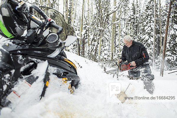 Älterer Mann im Ruhestand  der beim Schneemobilfahren mit der Kettensäge Wege räumt.