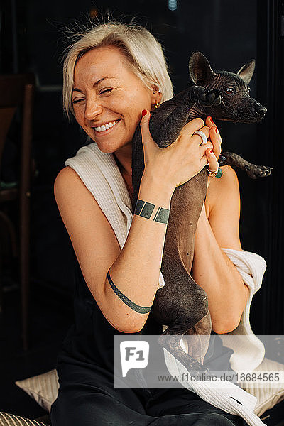 Blond woman holding dog in her hands while sitting and laughing