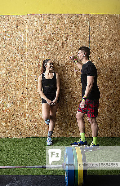 Strong man and woman in sportswear smiling and having nice conversation while leaning on gym wall