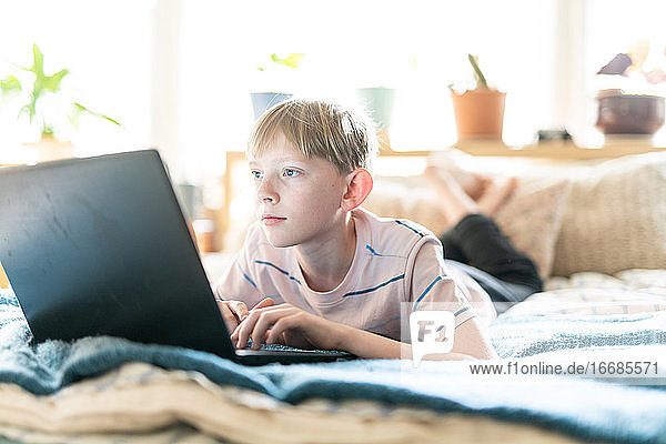 Teenager  der an einem Laptop arbeitet  während er in einem hell erleuchteten Raum auf dem Bett liegt