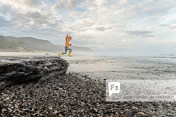 Junges Kind mit lockigem Haar springt von einem Felsen am Strand in Neuseeland