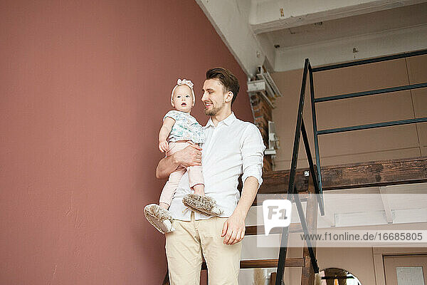 Ein Mann in einem weißen Hemd hält ein kleines Mädchen  das auf einer Treppe in seinem Haus steht.