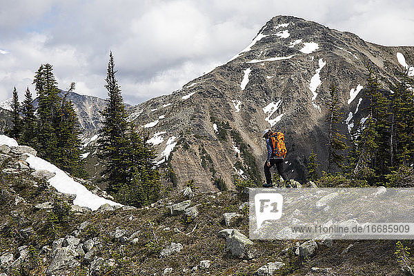 Side view of anonymous traveler with backpack hiking on stony slope of mountains covered with snow and forest in British Columbia in Canada