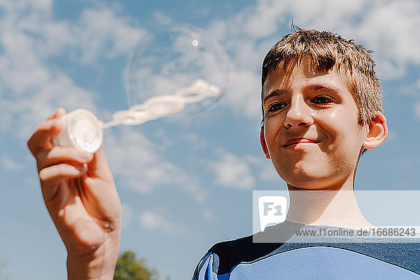 Junger weißer Junge  der eine Seifenblase im Freien mit Himmel im Hintergrund betrachtet