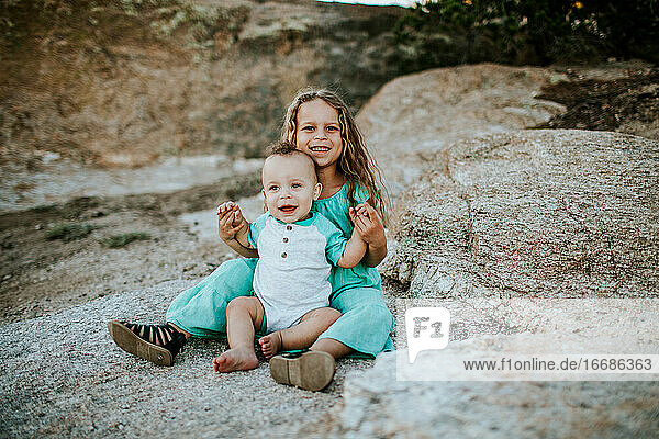 glückliches junges Mädchen  das seinen kleinen Bruder auf einem großen Felsen sitzt und hält