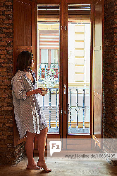 Junge Frau beim Frühstück neben einem Fenster zu Hause.