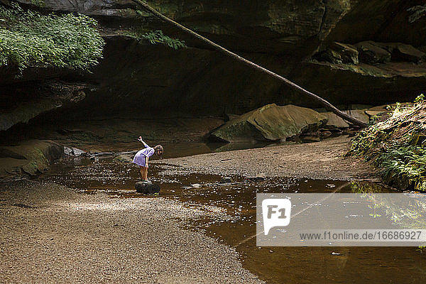 Ein kleines Mädchen steht auf einem Felsen und lehnt sich über den Bach  um die Spiegelung zu sehen