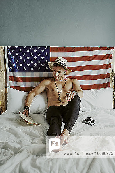 A young man in bed reading a shirtless book with the American fl