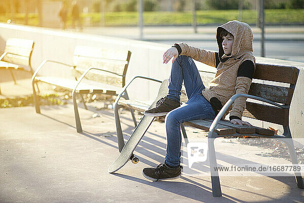 Junger Junge mit Kapuze sitzt auf einer Bank neben seinem Skateboard