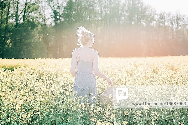 Frau steht in einem gelben Blumenfeld und hält einen Korb bei Sonnenuntergang