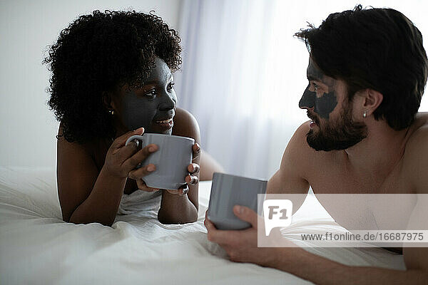 Multirassisches Paar mit Tonmasken auf dem Bett liegend