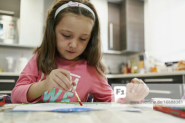Kleines Mädchen malt eine Zeichnung mit Aquarellfarben in ihrem Haus mit einer Küche im Hintergrund. Kreatives Konzept
