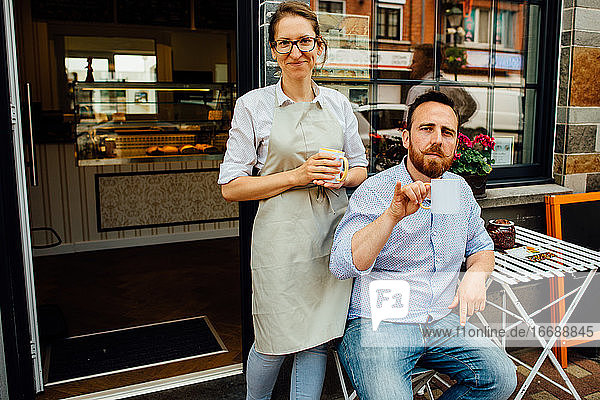 Mann und Frau lächeln in die Kamera  während sie am Eingang eines Cafés Tee trinken