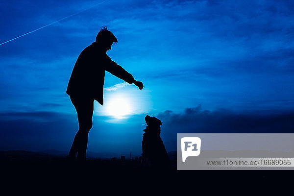Silhouette eines jungen Mannes  der seinen Hund mit blauem Nachtlicht füttert