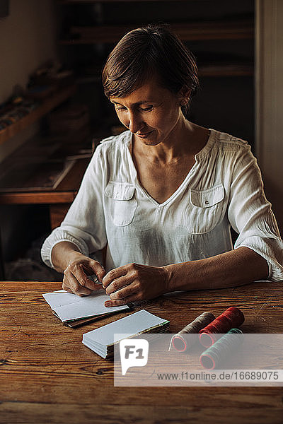 Am Tisch sitzende Kunsthandwerkerin  die Papierbögen für ein Buch näht