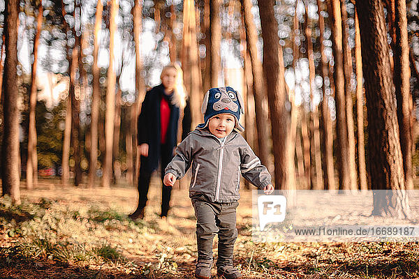 Baby boy walking in park in autumn with his mother standing behind him