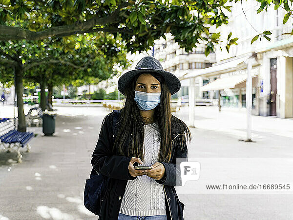 Junge Frau mit Maske  die in der Stadt ihr Mobiltelefon benutzt
