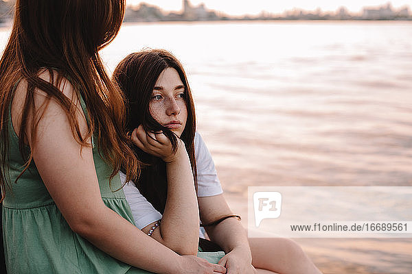 Thoughtful lesbian couple sitting by river in city at sunset