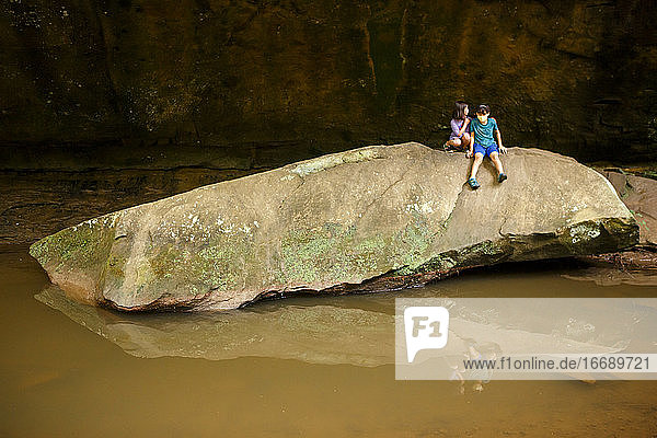 zwei Kinder sitzen auf einem großen Felsbrocken über einem spiegelnden Becken an einer Steinmauer