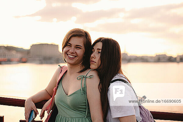Zwei glückliche Freundinnen stehen auf einer Brücke gegen den Fluss bei Sonnenuntergang