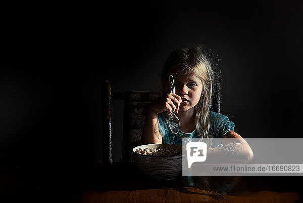 Kleines Mädchen isst Müsli in dramatischem Licht und schaut in die Kamera