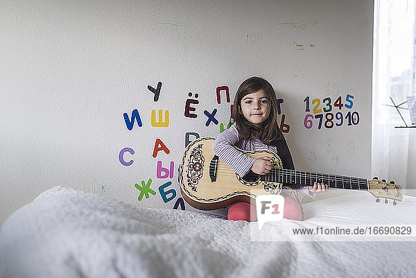 6-jähriges Mädchen mit Gitarre auf dem Bett vor einer Wand mit griechischem Alphabet