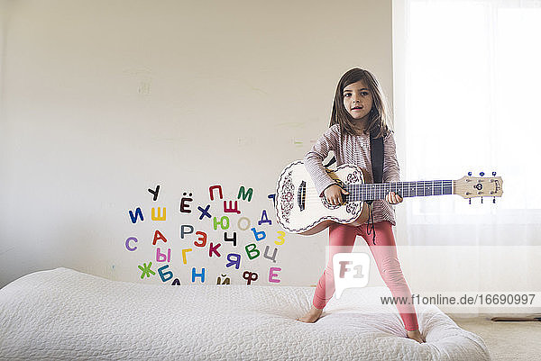 Junges Mädchen steht auf dem Bett und spielt Gitarre in einem Zimmer mit Buchstaben an der Wand
