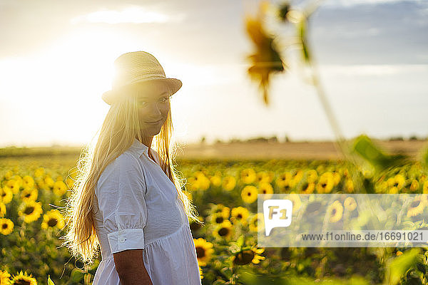 Junge attraktive blonde Frau posiert in ihrem Designerkleid in einem Sonnenblumenfeld und trägt einen Hut