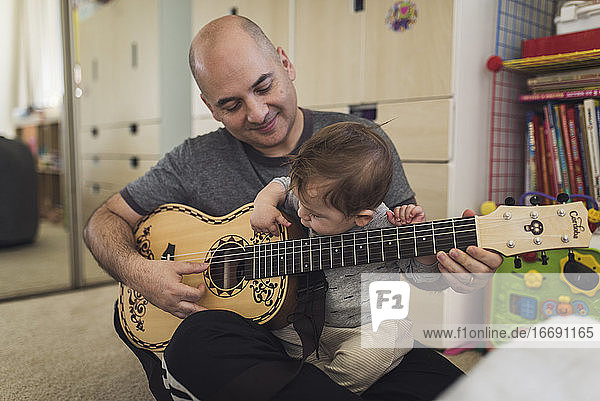 1 yr old playing child's guitar while sitting on bald father's lap