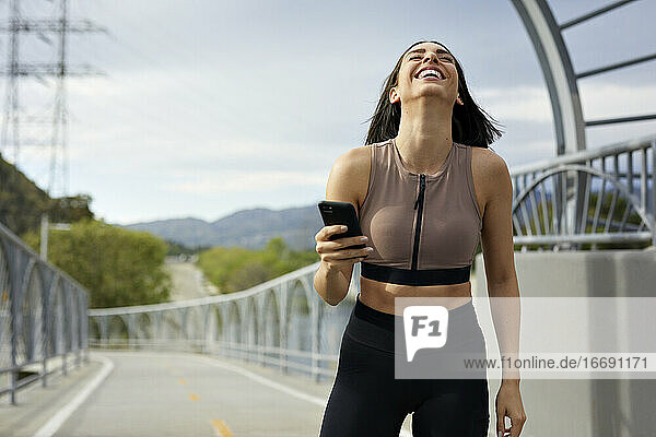 Fröhliche Frau in Sportkleidung mit Mobiltelefon auf einer Brücke stehend