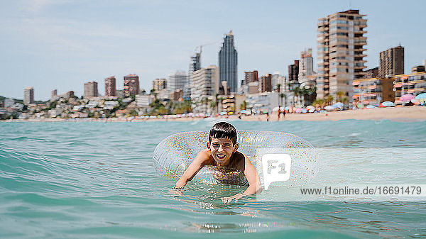 weißer Junge schwimmt in einem Schwimmer im Meer mit Gebäuden im Hintergrund