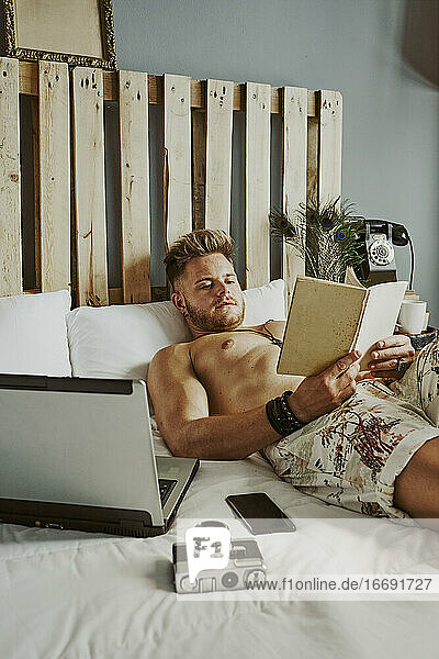 A man reads a book while working with his cell phone and laptop in a hotel bed